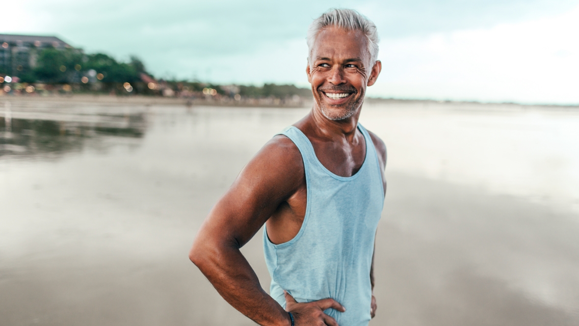 an older, tan, fit man stands on a beach and smiles