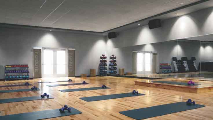 Grey yoga mats, each equipped with a pair of dumbbells, lined up on the gleaming wood floor of a mirrored studio  