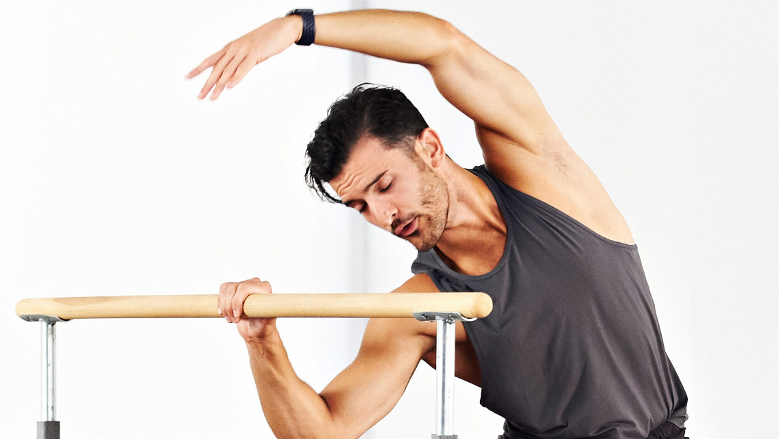 a man performs an overhead side reach while holding a barre