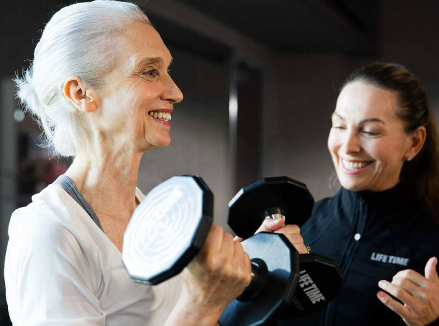 A Life Time personal trainer helps an woman on a piece of exercise equipment. 