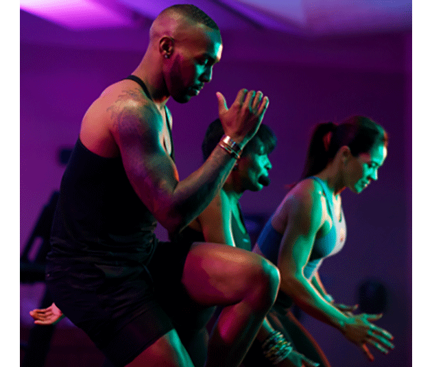  A side angle shot of a Black man from the knees up in an Ultra Fit class. He is exceptionally fit, with a full sleeve tattoo on his right arm, and is touching his right elbow to his left knee.