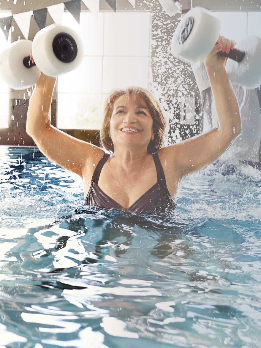 A woman lifts foam barbells over her head while in a pool.