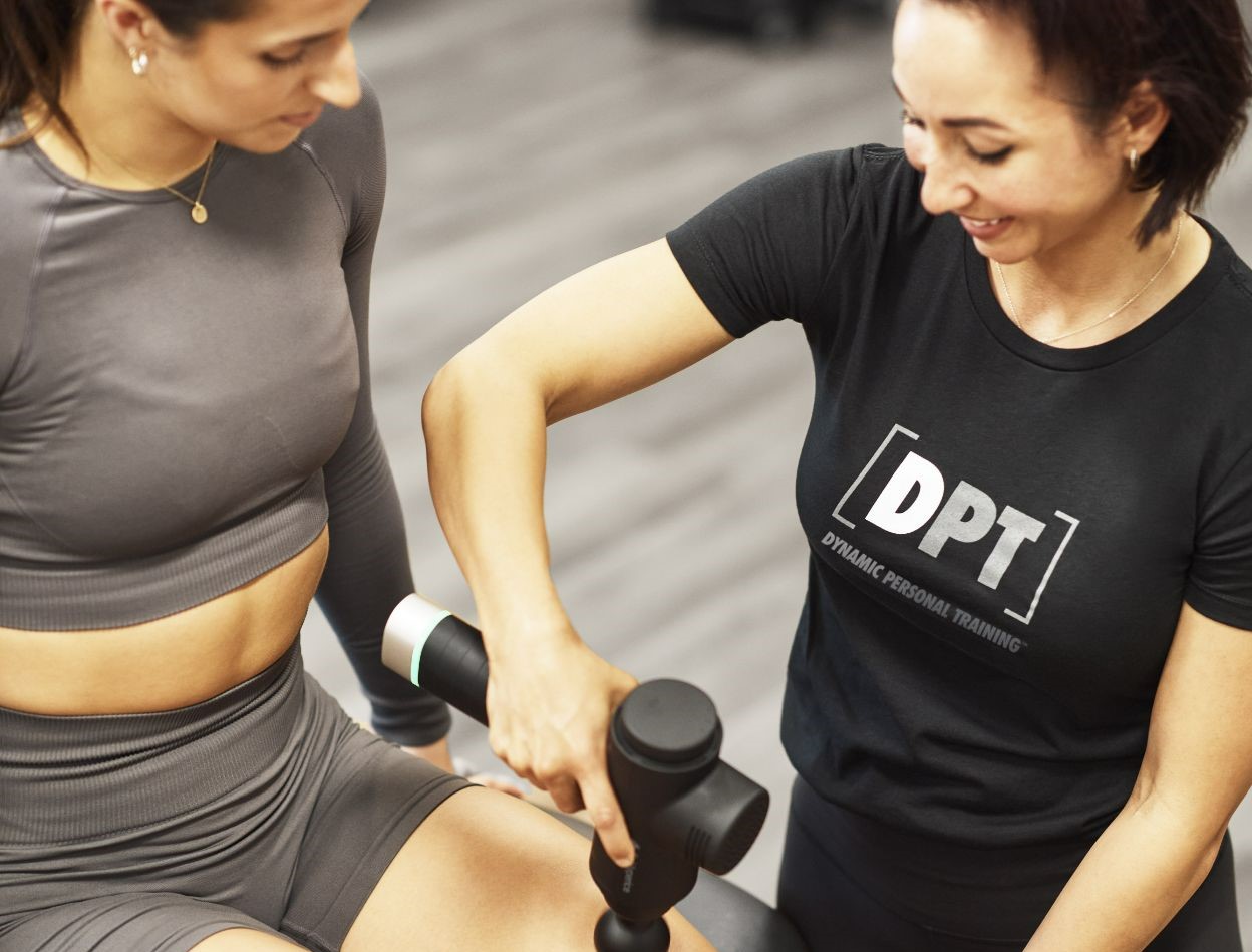 client sitting and relaxes while the trainer uses a massage gun on her quad 