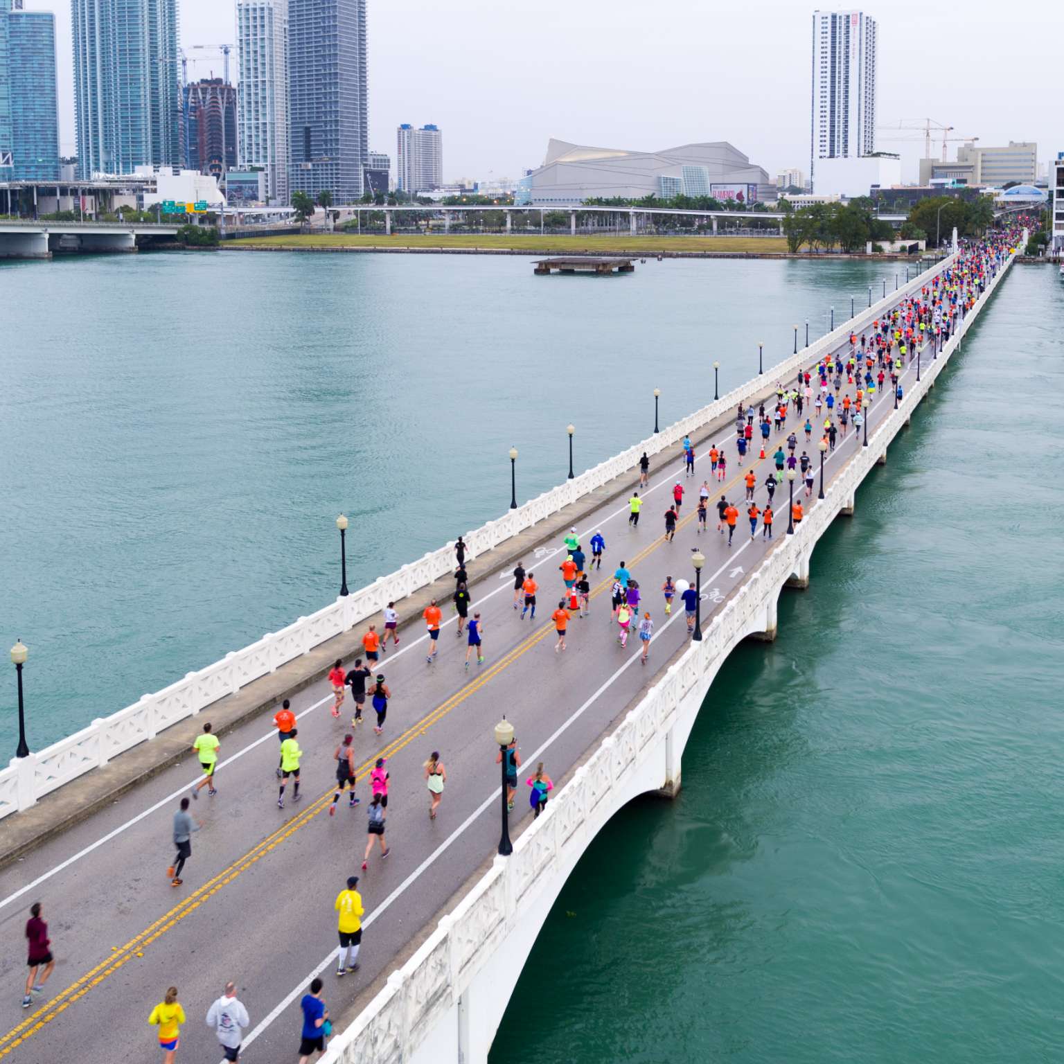 A crowd of runners running across a bridge over water with city buildings in the background