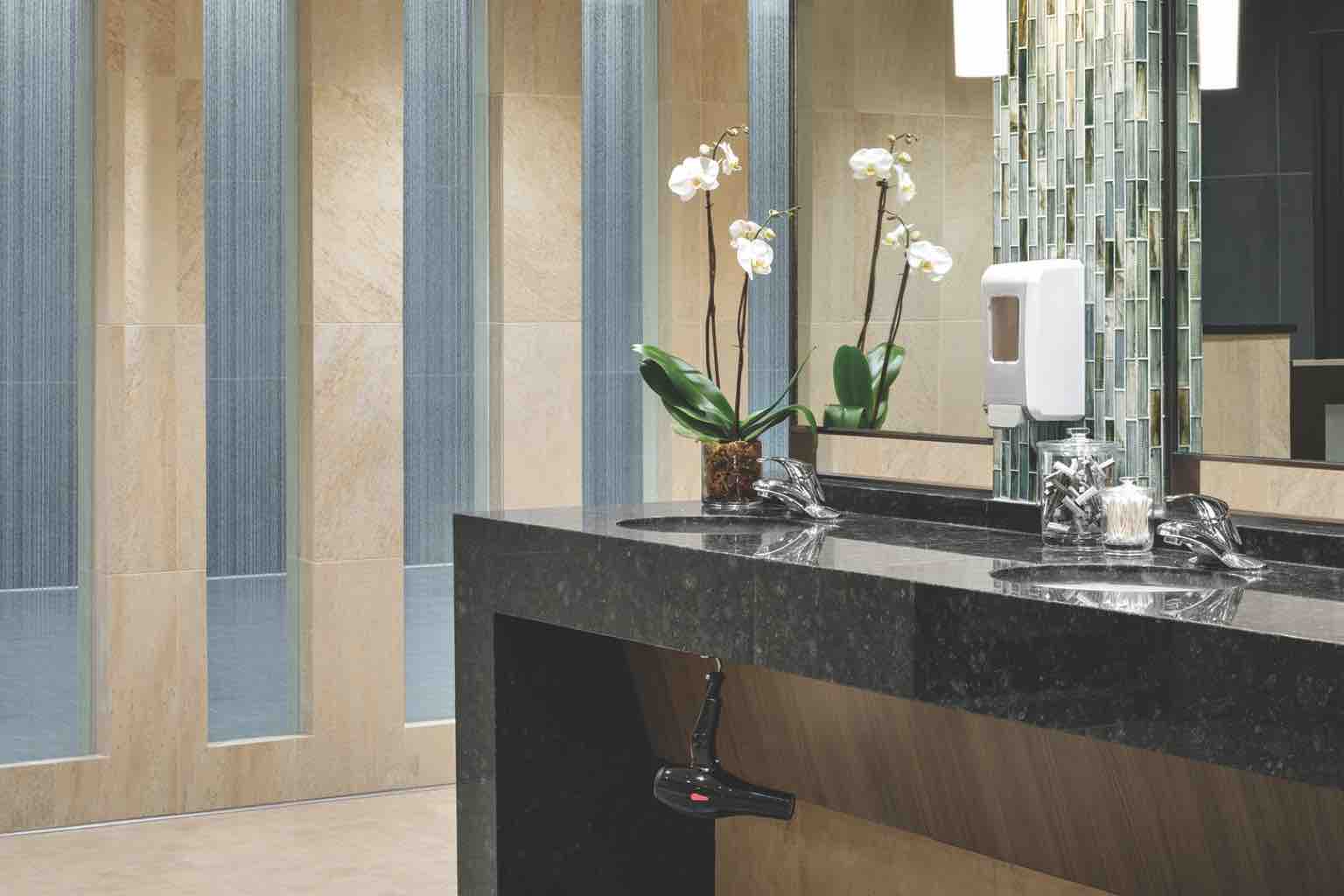 : A luxurious locker room with a row of three sinks in a black marble counter with tiled backsplash and hand soap dispensers