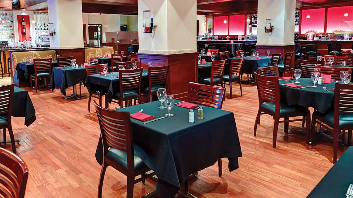 A wood-floored dining area with tables set with tablecloths, place settings, napkins and glasses