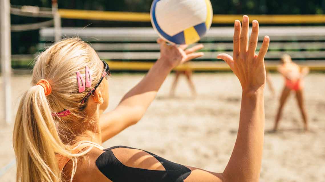 A woman sets up to serve a volleyball on an outdoor sand volleyball court