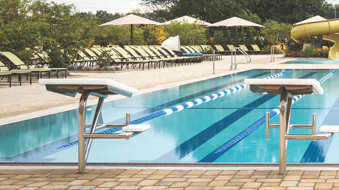 An outdoor pool deck at dusk, featuring a lap pool with starting platforms, chaises, umbrellas and a two-story waterslide