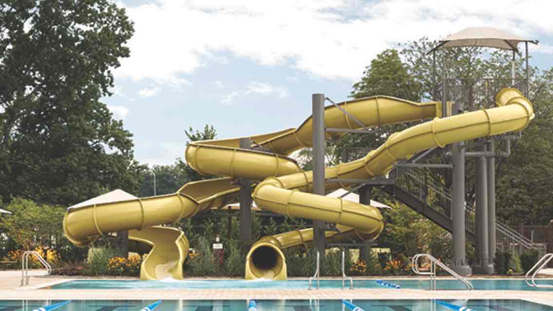 A large outdoor pool with two twisting yellow waterslides and green, leafy trees in the background