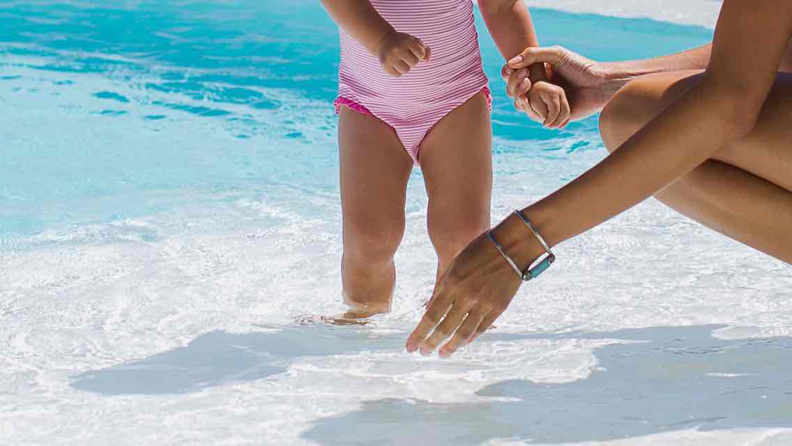 A little girl holds hands with her mom while they stand in ankle deep pool water