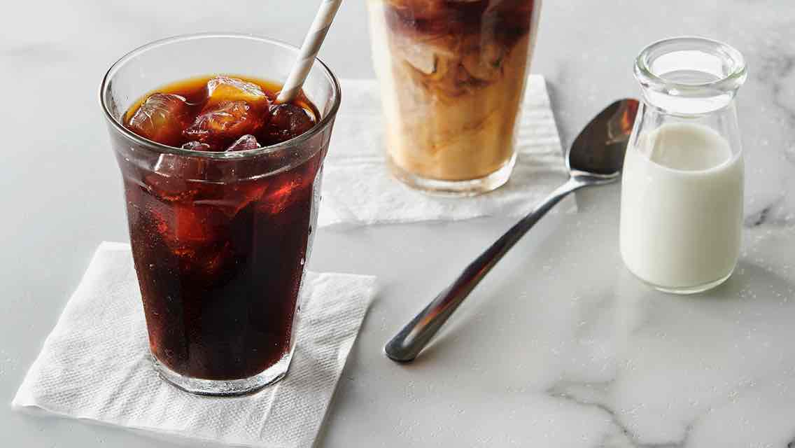 A gray and white marbled counter top with two glasses of iced coffee on white napkins, a metal stirring spoon and a small glass jar of milk