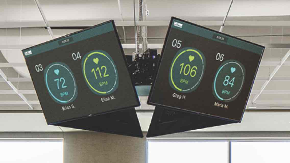 Two connected technology fitness monitors displayed near the ceiling in a studio class