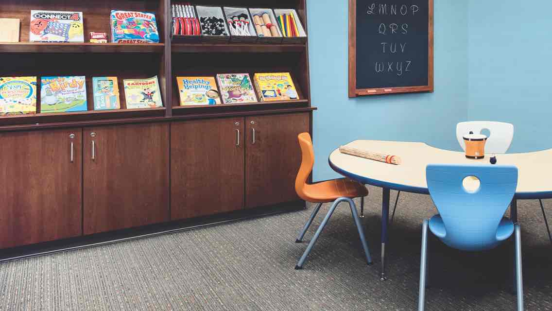 A children’s learning lab with wooden cabinets and shelves equipped with books, a small chalkboard on the wall and a table with three chairs