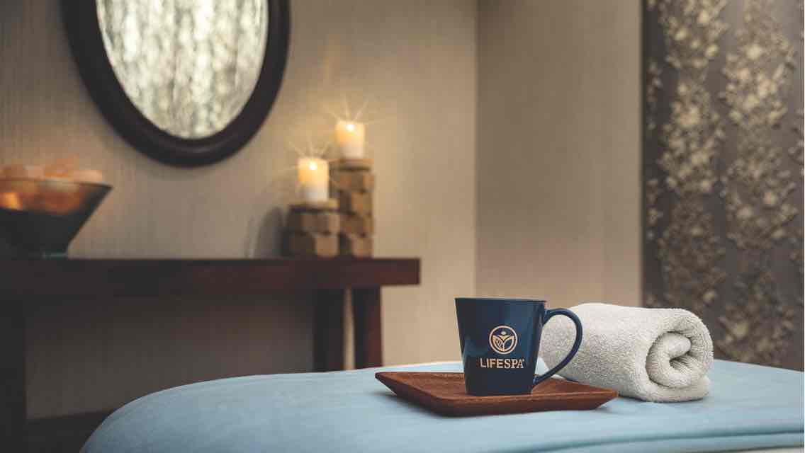 A rolled white towel and a mug with a LifeSpa logo sit atop a blanket-covered massage table 