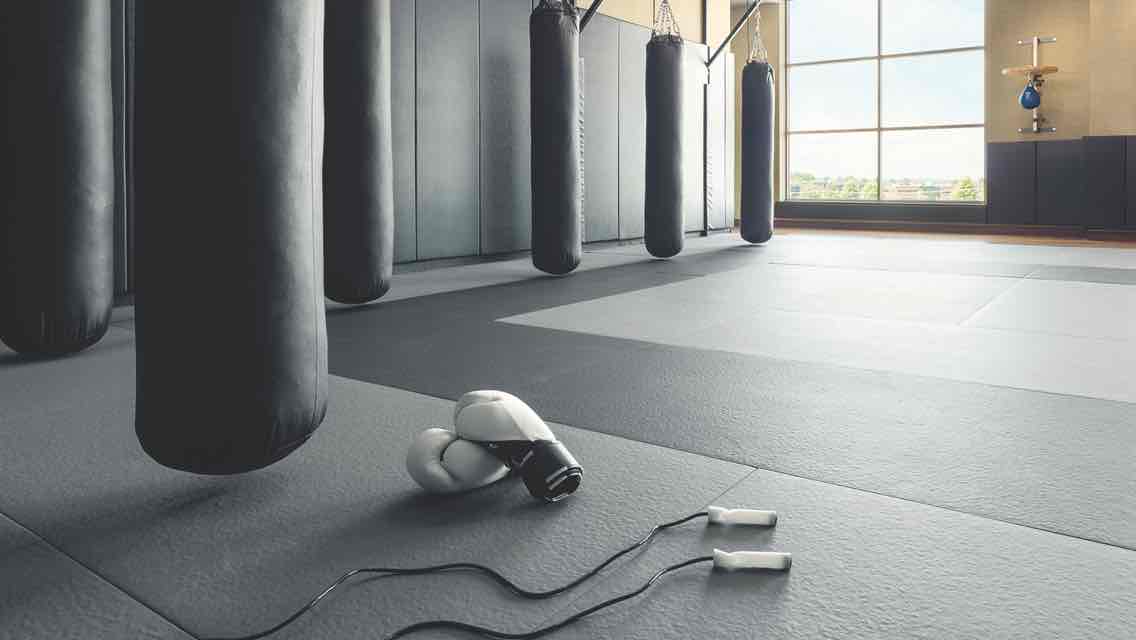 A sunlit boxing gym with a padded gray and black floor and a row of six black hanging punching bags  