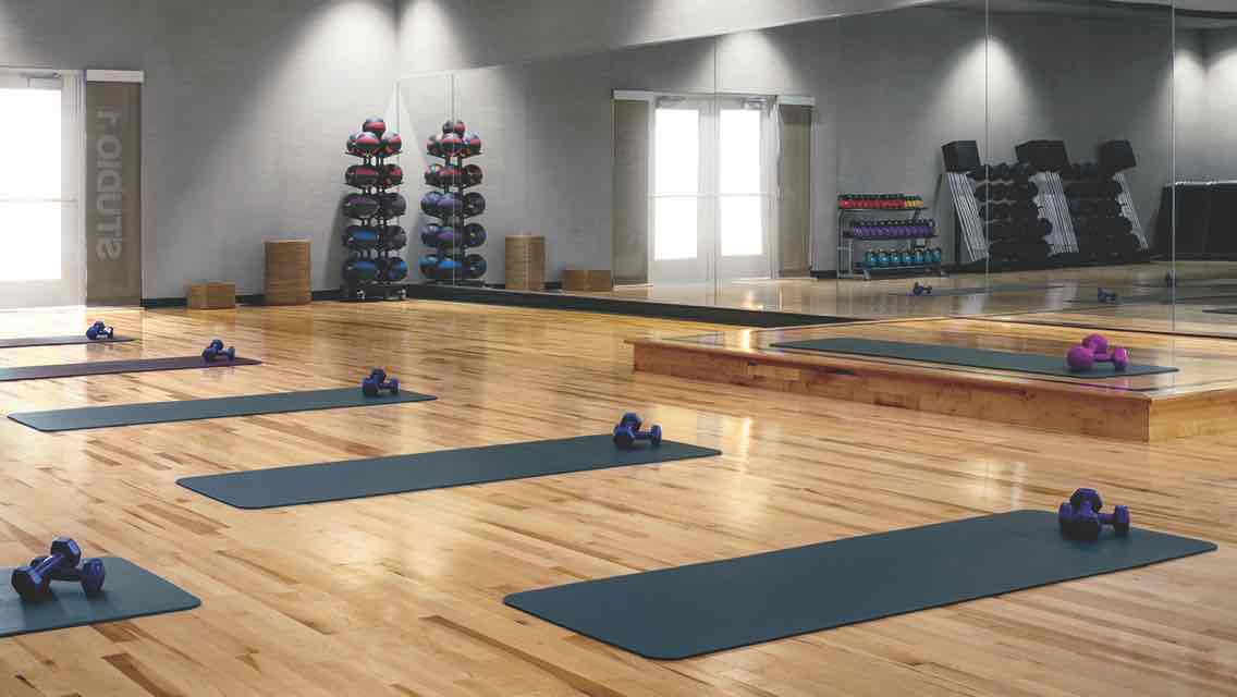 Blue yoga mats, each equipped with a set of hand weights, lined up on a studio’s wood floor