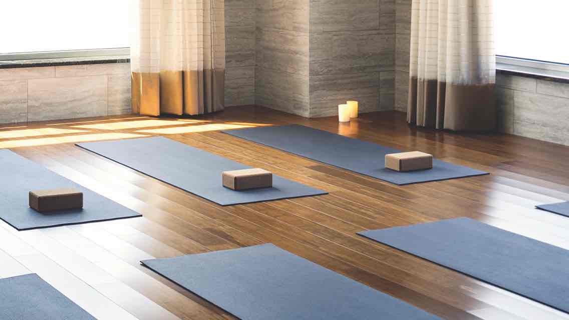 Blue yoga mats, each equipped with a yoga brick, lined up on the wood floor of a candlelit studio