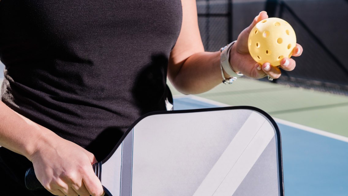 Person holding pickleball and pickleball paddle on a tennis court. 