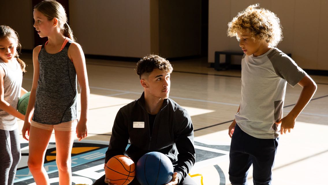 A sports coach holds two basketballs while instructing three children