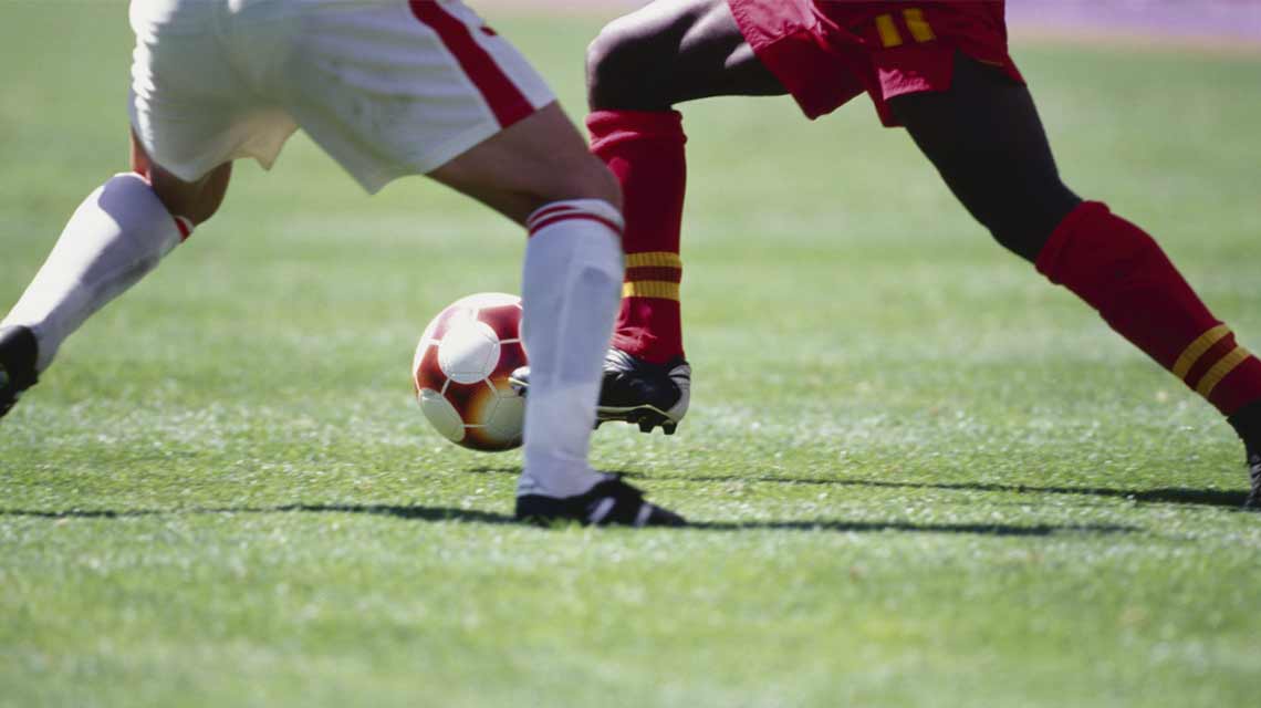 The legs of 2 soccer players as one dressed in a red uniform dribbles the soccer ball past the one dressed in a white uniform