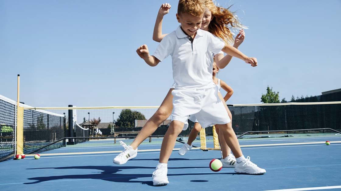 A young girl and young boy dance over a tennis ball on an outdoor tennis court