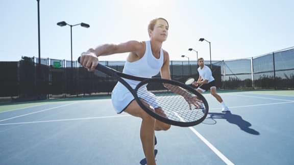 A woman in a white tank top and tennis skirt steps forward on an outdoor tennis court to take a one-handed backhand swing with a tennis racquet