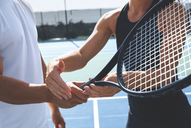 A female tennis instructor in a black tennis outfit helps a male student in a white tennis outfit perfect his grip on a tennis racquet on an outdoor tennis court