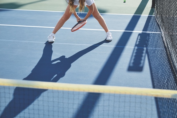 A girl in white tennis shorts stands in the ready position and holds a tennis racquet on an outdoor tennis court.