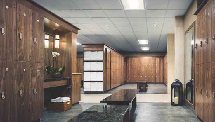 A large locker room with tiled floors, wooden lockers, gleaming benches and shelves filled with neatly folded white towels. 