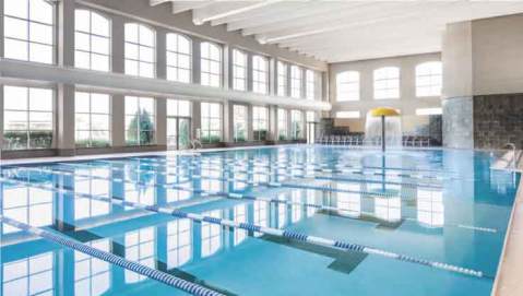 A wall of windows facing a large indoor pool with blue and white lane dividers and a yellow mushroom-shaped spray fountain