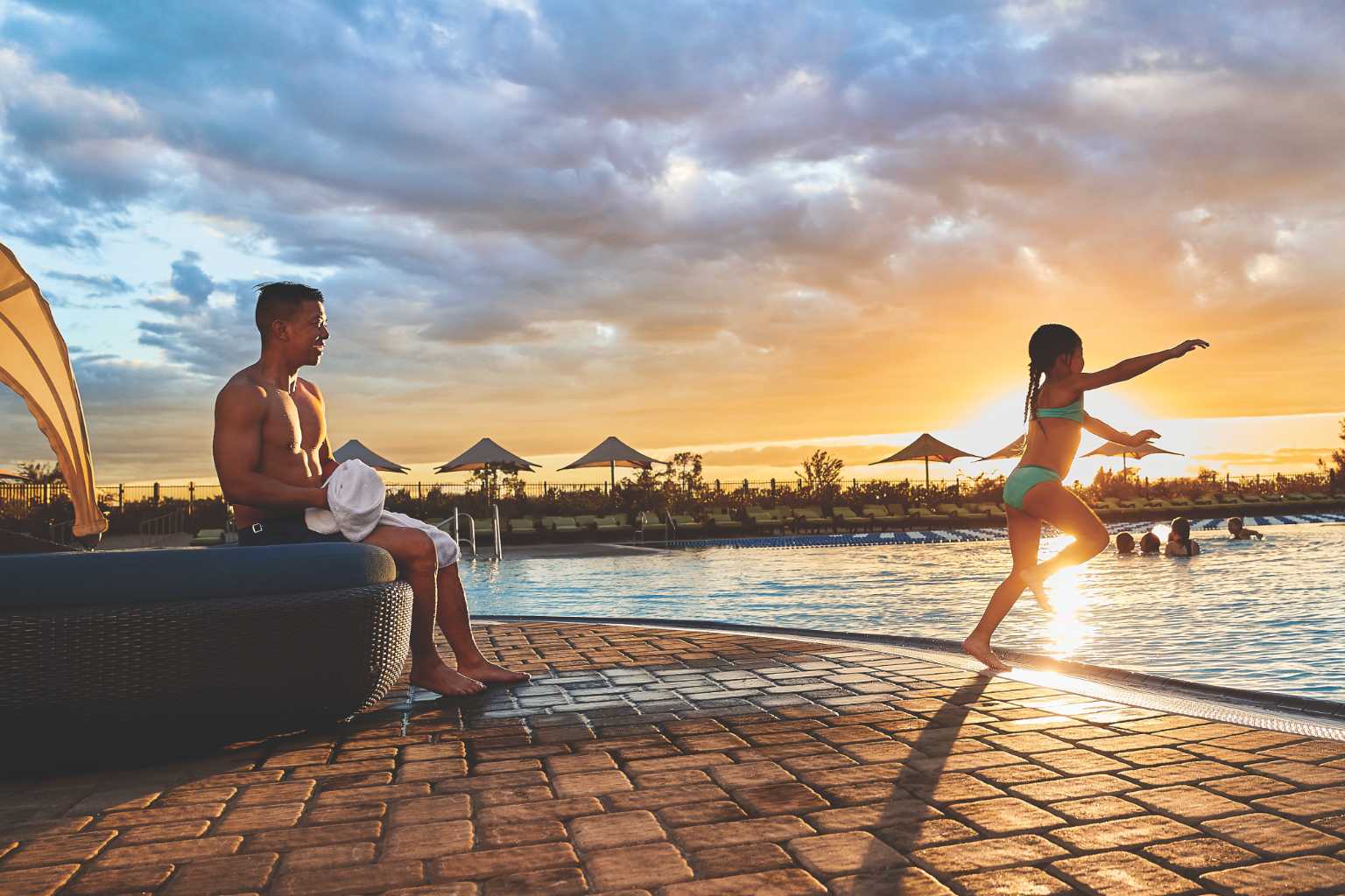 Girl jumping into a pool at sunset with her dad in a lounge chair behind her.
