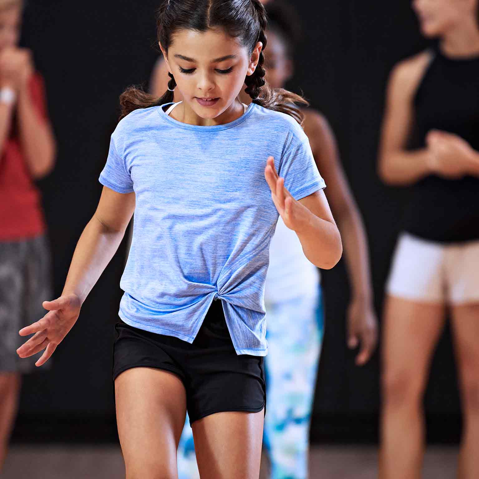 A waist up shot of a girl wearing a light blue t-shirt with brown hair pulled back into two long braids. The looks down at the floor, completely focused on the drill she is doing. Other kids and an adult are visible in the background.