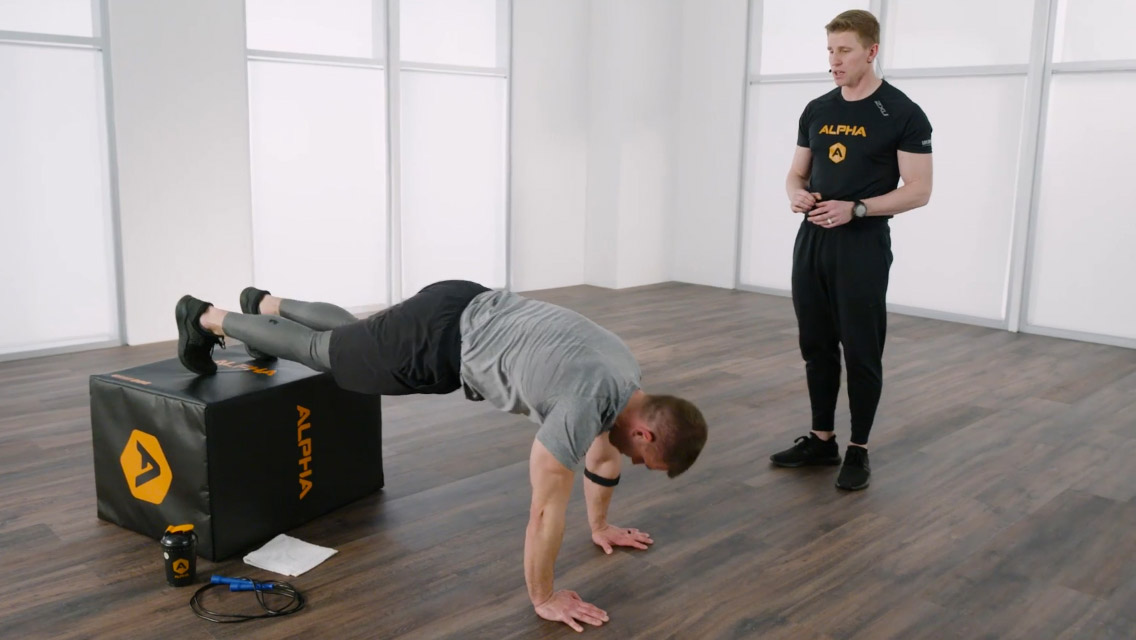 A man performs push-ups with his feet propped on an Alpha box  