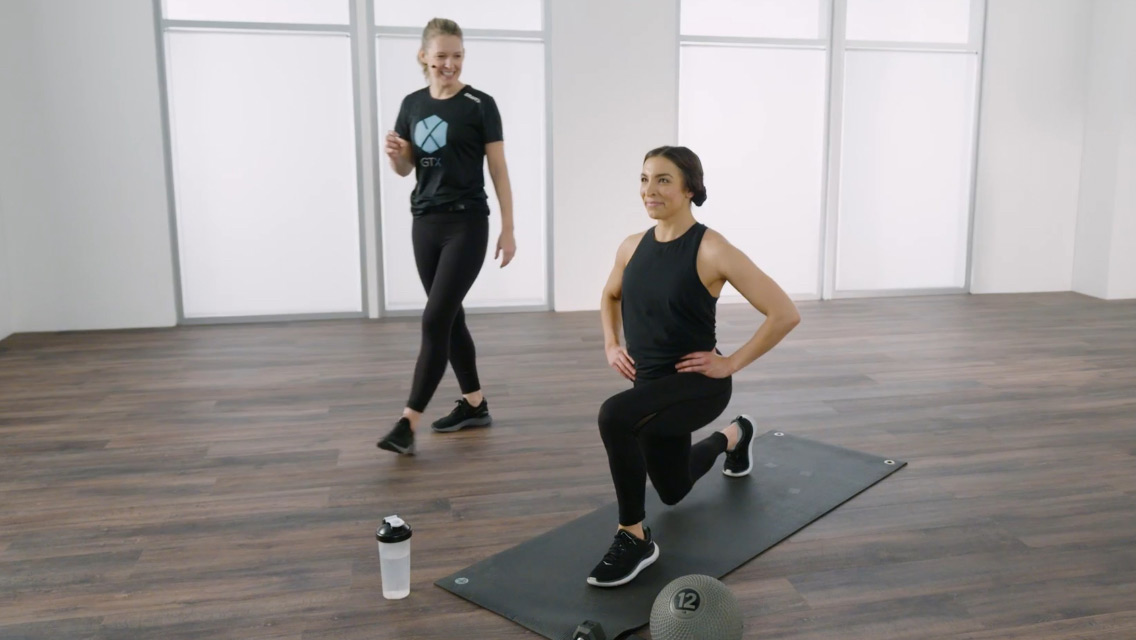 A student on a yoga mat performs a lunge under the supervision of  a Life Time Trainer 