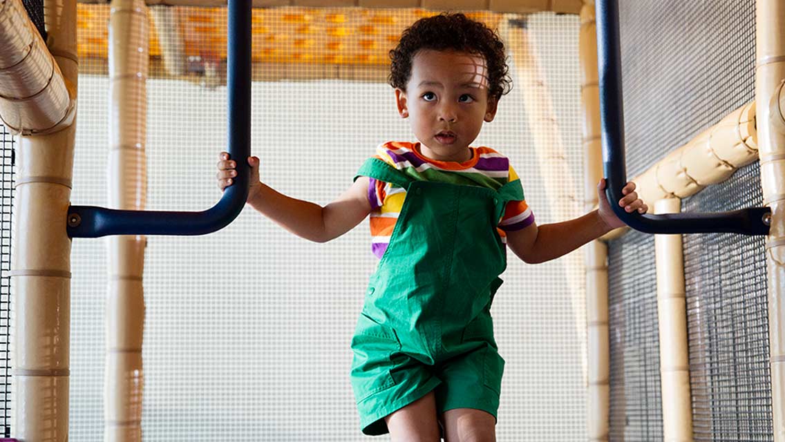 A child in green overalls playing on an indoor jungle gym