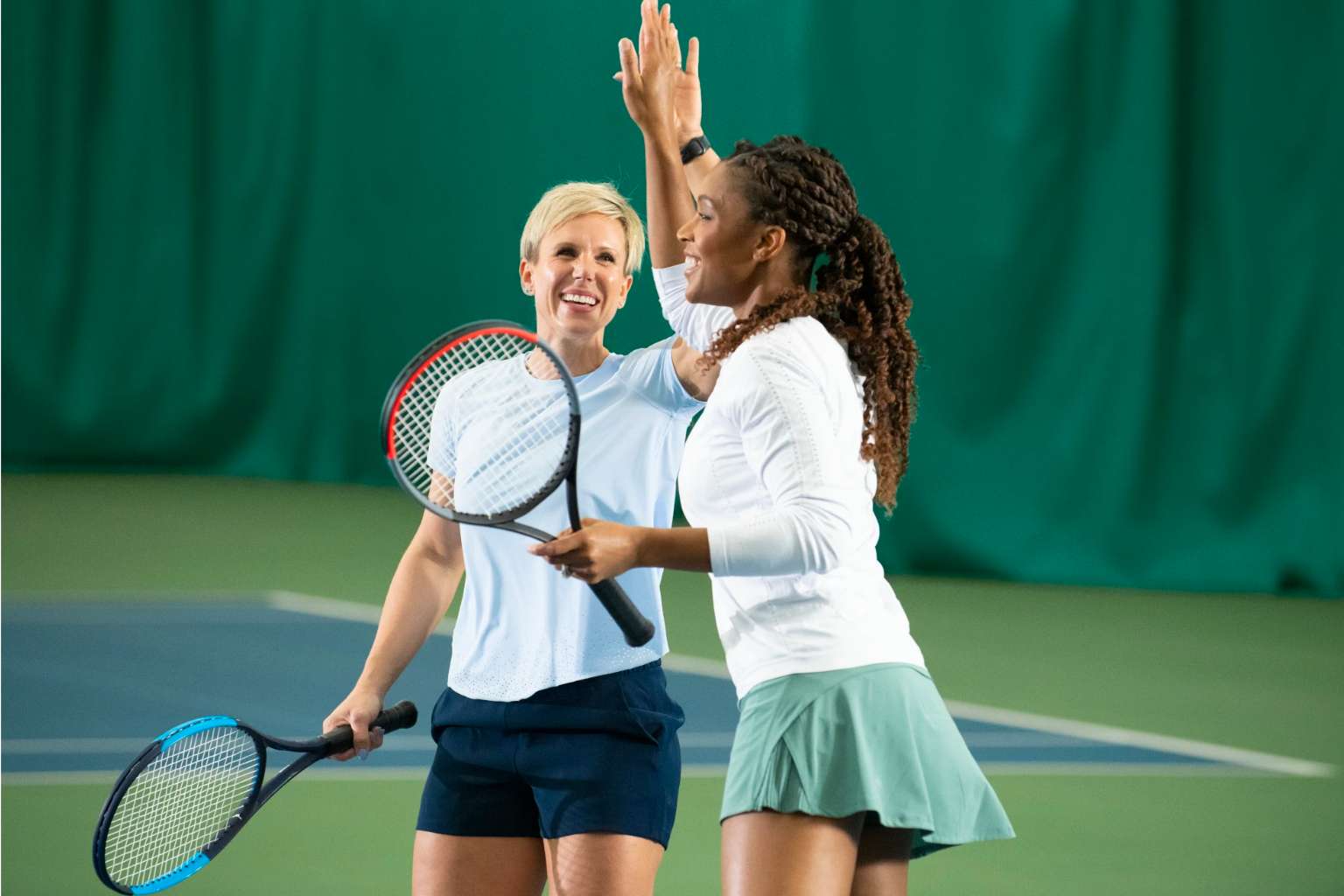 Two smiling women holding racquets and dressed in tennis gear high five on an indoor court