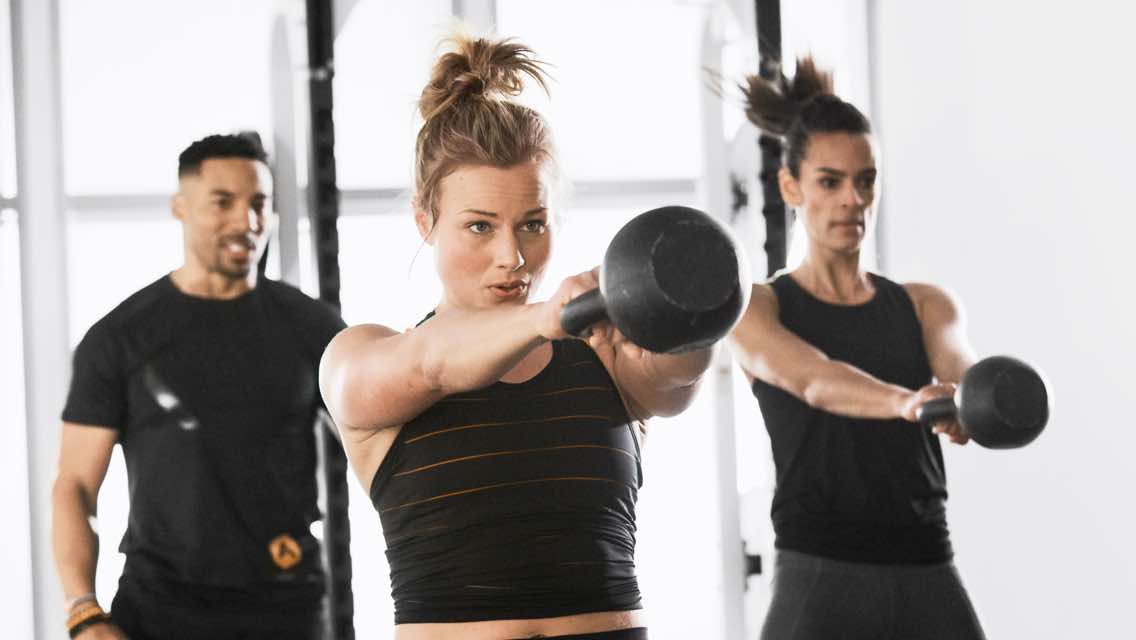 Two women working out kettlebells while a trainer cheers them on