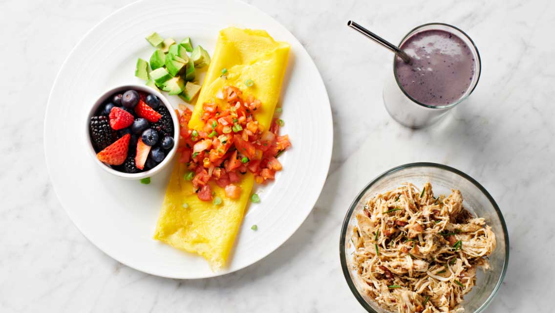 An over head view of a plate with an omelet, salsa and fruit; a bowl of shredded, seasoned chicken; and a purple smoothie.