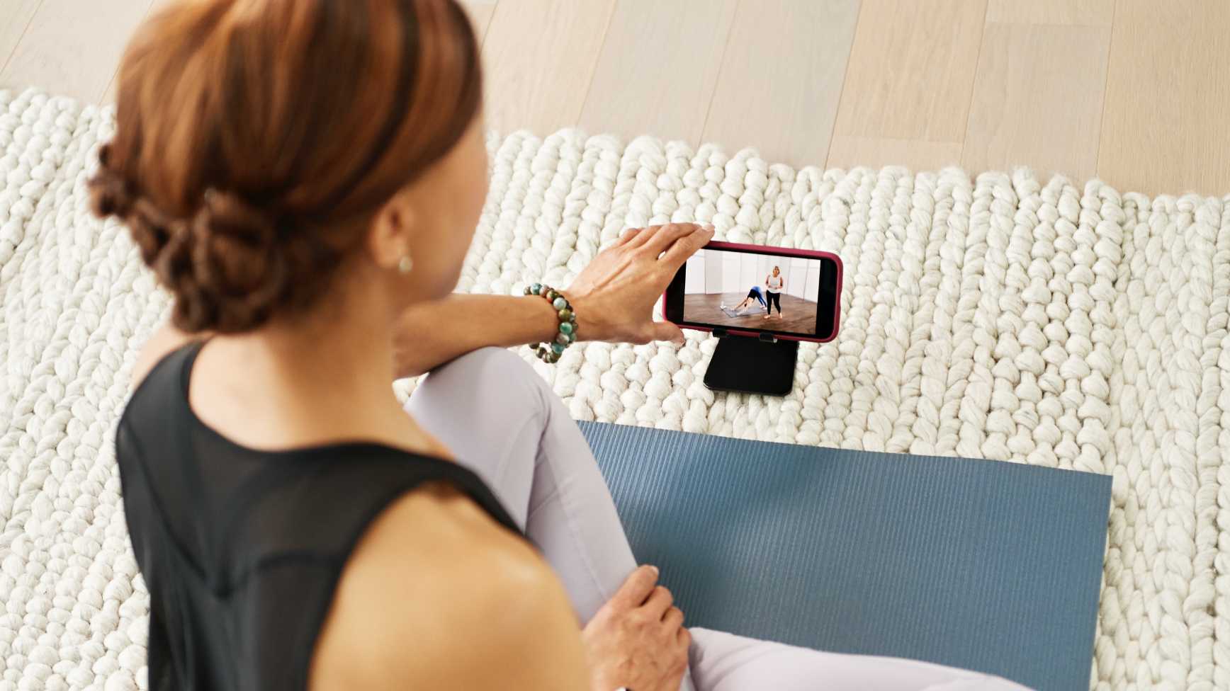 Woman in living room sitting on yoga mat, watching a workout class on her phone.