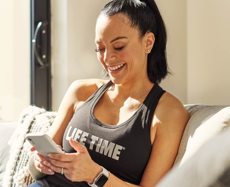 A smiling woman wearing a black tank top, looks at her phone while sitting on her couch. 