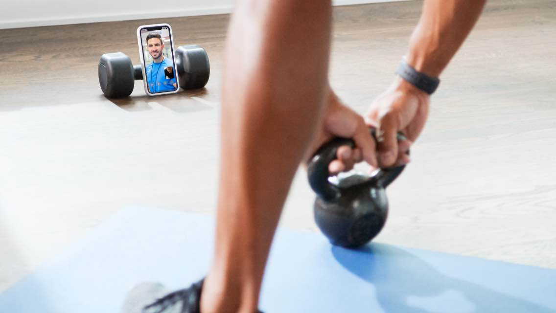 A close up shot of a phone, propped up against a dumbbell so that a personal trainer can watch him work out. 