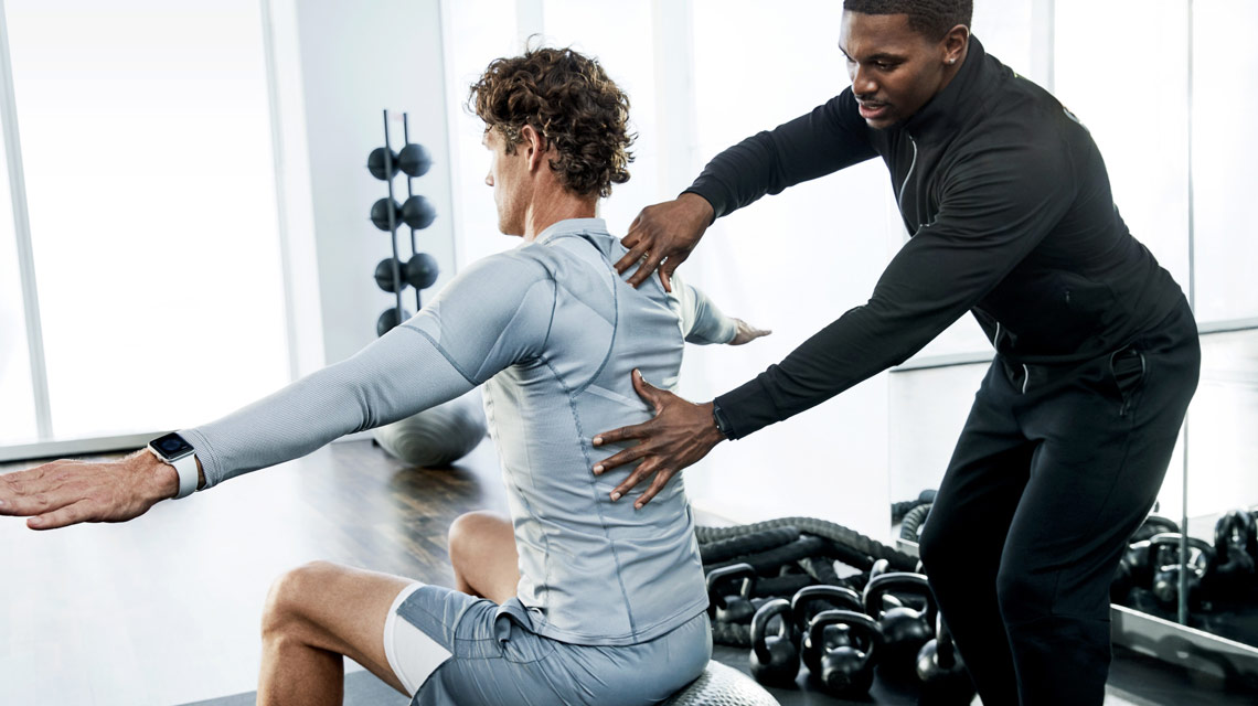 a therapist placing his hands on the back of a client who is sitting at a bench with his arms out, surrounded by free weights