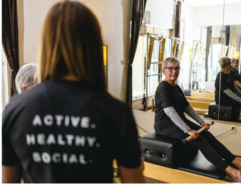 a woman sits on a pilates reformer machine and smiles at her instructor