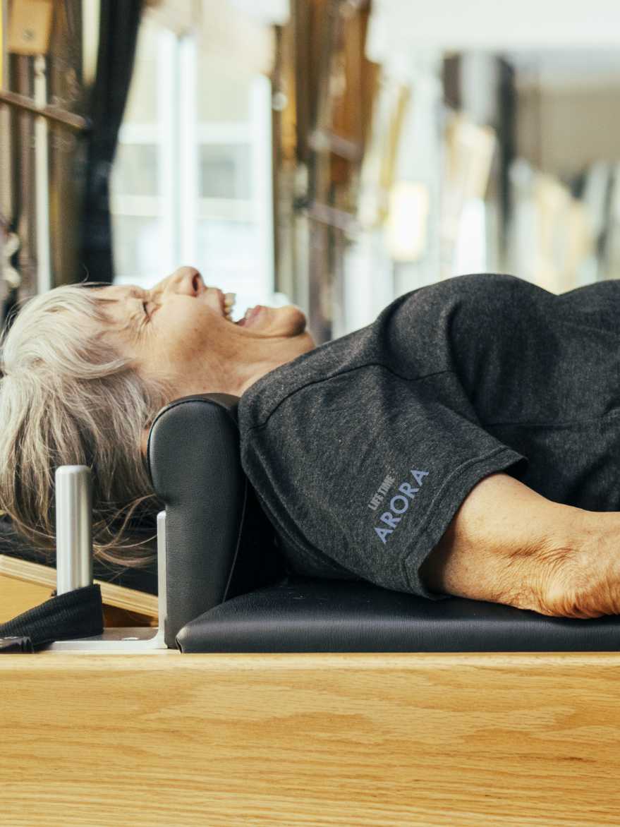 A woman lies on her back on a pilates reformer machine and performs a pilates exercise.